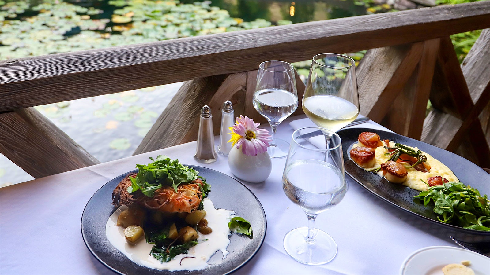 A table at Mirbeau Inn & Spa near a pond and a bridge, with glasses of water and wine and plates of fish and scallops